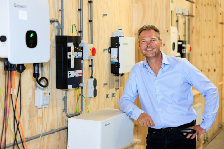 Het beeld toont een man in een blauwe shirt die ontspannen poseert naast een opstelling van elektrische apparatuur tegen een houten wand. De installatie bestaat onder andere uit zonnepanelenomvormers, bevestigd op een efficiënte en georganiseerde manier. De setting is waarschijnlijk in een technische omgeving waar duurzame energie centraal staat. De foto is gemaakt door Jan Buwalda in Groningen.