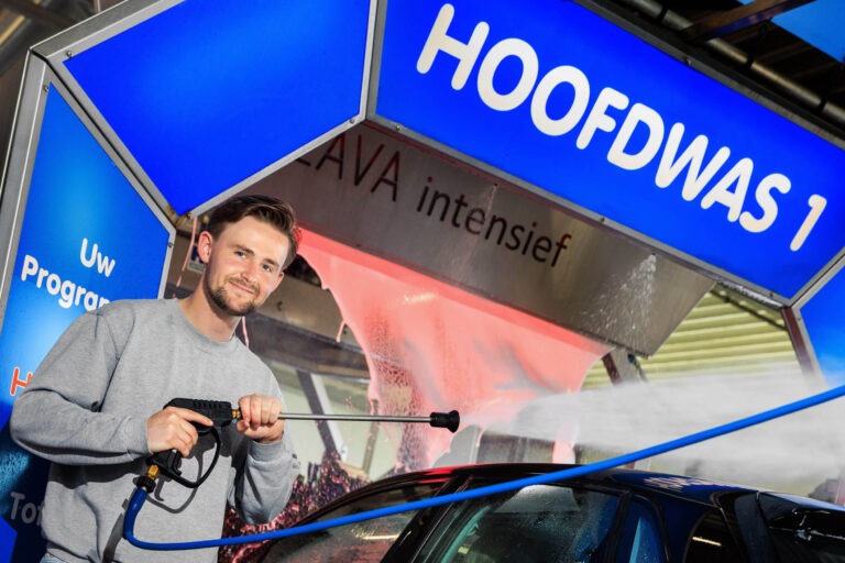 Een man staat met een hogedrukspuit bij een wasstraat, klaar om een auto schoon te maken. Hij draagt een grijze trui en glimlacht naar de camera. Op de achtergrond is een deel van de wasstraat te zien met het bord HOOFDWAS 1. De omgeving is helder verlicht en er is schuim zichtbaar op de auto. Foto door Jan Buwalda uit Groningen.