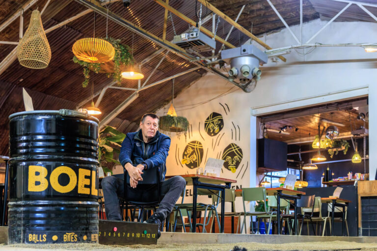 Dit beeld toont een man zittend in een sfeervolle ruimte met een industriële uitstraling. De ruimte heeft een houten plafond en decoratieve hanglampen, met planten en moderne kunst aan de muur. Op de voorgrond staat een zwarte ton met het opschrift BOEL. De sfeer is uitnodigend en informeel, passend bij een recreatieve ruimte. Fotograaf: Jan Buwalda, Groningen.