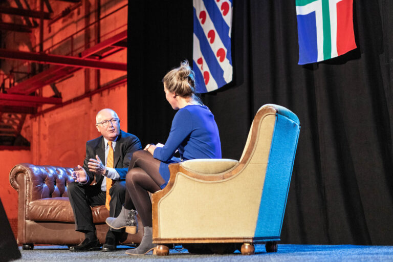 Een man en een vrouw zitten tegenover elkaar op een podium tijdens een gesprek. De man draagt een donker pak met een oranje stropdas en zit op een bruine leren bank. De vrouw draagt een blauwe jurk en zit op een beige stoel met blauwe accenten. Achter hen hangen een Friese en een Zuid-Afrikaanse vlag. Op de achtergrond is een industriële muur te zien, verlicht met warme, oranje tinten. Foto door Jan Buwalda, Groningen.