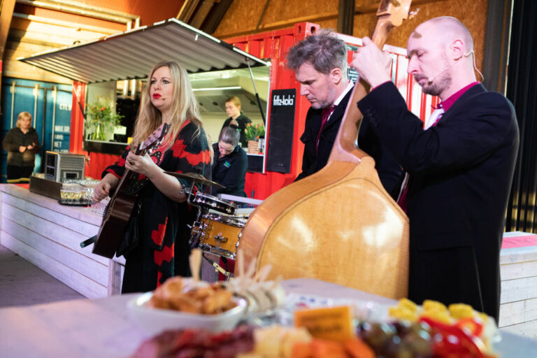 Ik zie een muziekoptreden met een vrouw die gitaar speelt en twee mannen die respectievelijk een contrabas en drumstel bespelen. Ze staan op een podium voor een rood containerachtige structuur. Op de voorgrond zijn er diverse hapjes gepresenteerd. De foto is levendig en toont een gezellige sfeer. Fotograaf: Jan Buwalda, Groningen.