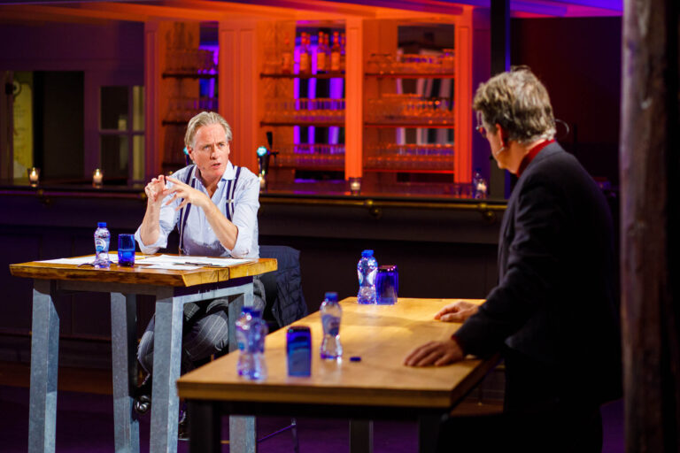 Dit is een foto waar twee mannen betrokken lijken in een gesprek aan een houten tafel. De ruimte heeft een sfeervol verlichte bar op de achtergrond met warme kleuren. Er staan flessen water op de tafels. De setting lijkt professioneel en ongedwongen. Fotografie door Jan Buwalda in Groningen.