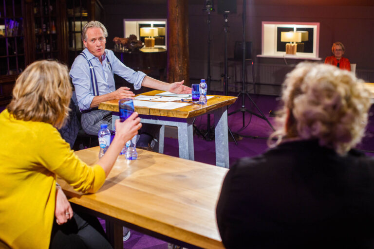 Een man zit aan een tafel in een ruimte met een paarse vloer en houten meubilair. Hij draagt een lichtblauw overhemd en bretels, en er liggen papieren en pennen voor hem. Er zijn andere mensen aan tafels te zien, waarvan één persoon een blauw glas vasthoudt en een ander zich richt op de spreker. De sfeer lijkt informeel en professioneel. Fotograaf: Jan Buwalda, Groningen.