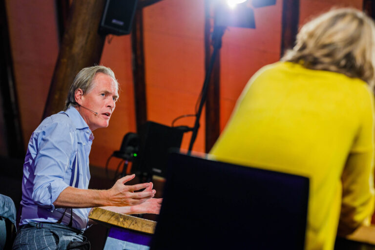 Dit beeld toont een man gekleed in een lichtblauw overhemd die een gesprek voert met een andere persoon, mogelijk tijdens een discussie of presentatie. Hij draagt een microfoon aan zijn oor en is levendig aan het gebaren, wat kan duiden op een geanimeerd gesprek of presentatie. De achtergrond is sfeervol verlicht met warme tinten, en er is een felle lamp zichtbaar, die de man vanuit de zijkant belicht. De andere persoon in het beeld draagt een felgele top, maar staat met de rug naar de camera. Deze foto is gemaakt door Jan Buwalda in Groningen.