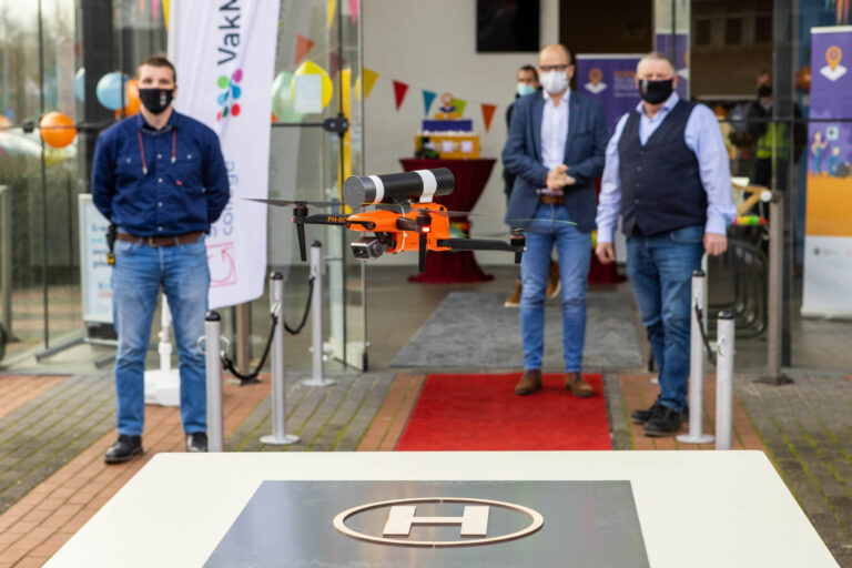 Een middelgrote oranje drone vliegt boven een landingsplatform met daarop een 'H'. De drone bevindt zich voor de ingang van een gebouw, geflankeerd door meerdere mannen die allen mondkapjes dragen. Op de achtergrond zijn kleurrijke ballonnen en vlaggenlijnen te zien, wat een feestelijke sfeer suggereert. De foto is gemaakt door Jan Buwalda in Groningen.