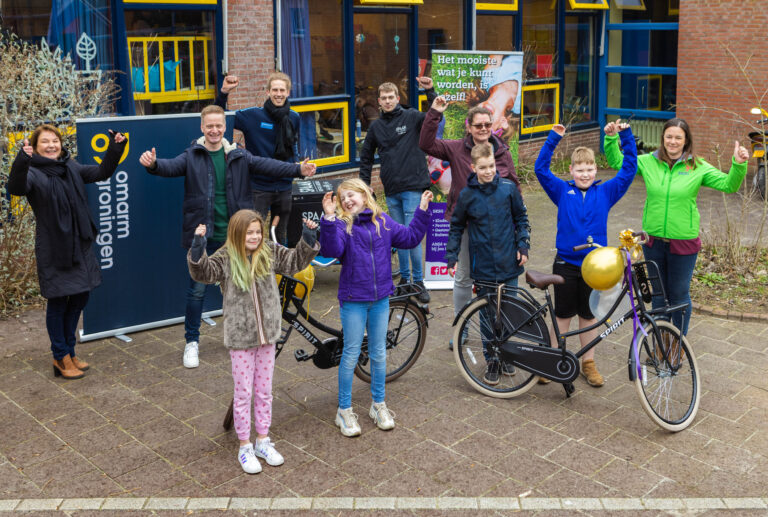 Op deze foto zien we een groep enthousiaste mensen die met opgeheven armen poseren bij een banner van Omatrin Groningen. Er zijn zowel volwassenen als kinderen aanwezig, glimlachend en blij. Er staat ook een fiets met een grote gouden ballon erop. De sfeer is vrolijk en feestelijk. De foto is gemaakt door Jan Buwalda in Groningen.
