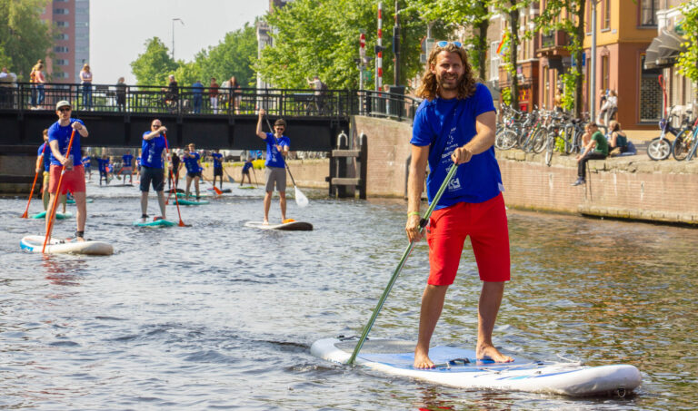 Op de foto van Jan Buwalda zien we een groep mensen die aan het suppen is in de grachten van Groningen. De deelnemers, gekleed in blauwe T-shirts, peddelen ontspannen over het water op een zonnige dag. Op de achtergrond is een brug te zien met toeschouwers die toekijken. De historische stad en het groene gebladerte langs de kade geven een gezellige en levendige sfeer aan de scene.