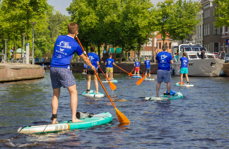 Op een zonnige dag in Groningen zien we een groep mensen die aan het suppen zijn op de grachten. Ze dragen blauwe T-shirts en peddelen rustig door het water, omringd door groene bomen en typische Nederlandse architectuur. De sfeer is ontspannen en gezellig. Foto door Jan Buwalda.