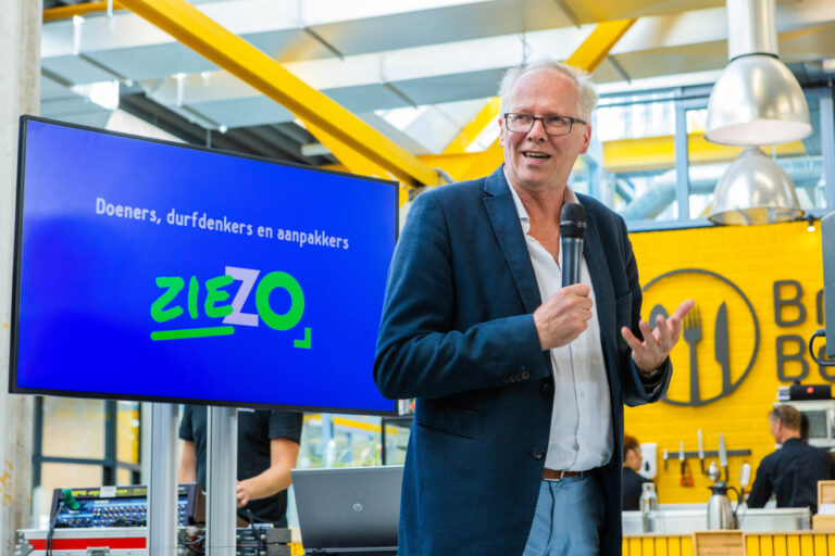 Dit is een foto van een man die in een moderne ruimte een presentatie geeft. Hij draagt een donkerblauw colbert en een witte overhemd, en spreekt in een microfoon. Achter hem is een groot scherm te zien met de tekst Doeners, durfdenkers en aanpakkers en het logo ZIEZO. De ruimte heeft een industriële uitstraling met gele accenten en een logo van bestek op de muur. Fotograaf: Jan Buwalda, Groningen.