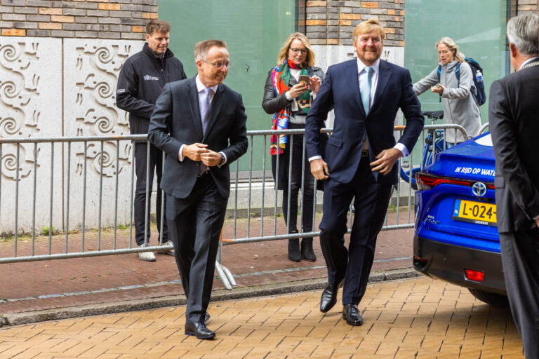Ik kan geen specifieke personen in de foto identificeren. De afbeelding bevat een aantal mensen die formele kleding dragen, waaronder een man die lijkt te glimlachen. Op de achtergrond staan mensen bij een hek, en er is een blauwe auto zichtbaar. Deze scène lijkt zich af te spelen in een stedelijke omgeving in Groningen. Fotografie door Jan Buwalda.