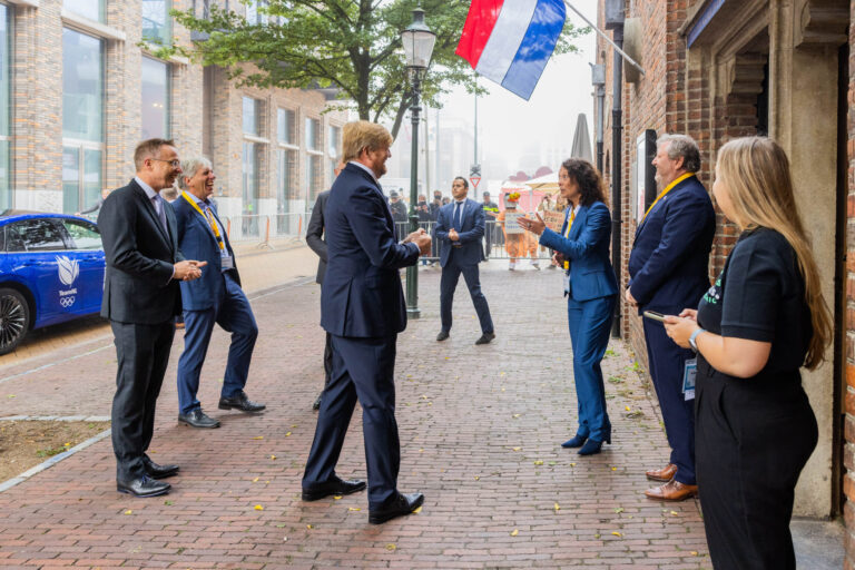 Het beeld toont een groep mensen die op een straat in Groningen staan, gekleed in nette kleding. Ze lijken in een gesprek of discussie verwikkeld te zijn. Er hangt een Nederlandse vlag boven hen. Op de achtergrond is een auto met het Team NL-logo te zien. De sfeer oogt professioneel en vriendelijk. Fotograaf: Jan Buwalda.