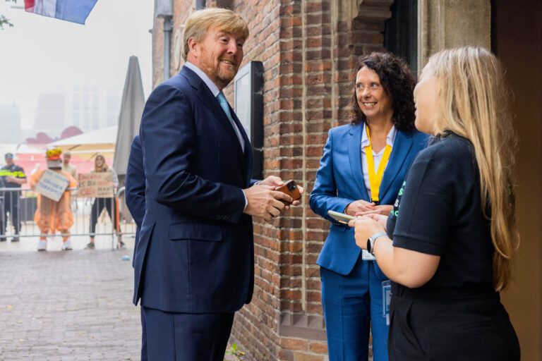 Het beeld toont een man in een donkerblauw pak die vriendelijk in gesprek is met twee vrouwen. Een van de vrouwen draagt een blauw pak en een gele lanyard om haar nek, terwijl de andere vrouw gekleed is in een zwart shirt. Op de achtergrond zijn protesterende mensen te zien met borden, en een politieagent staat in de buurt. De scène speelt zich af buiten, naast een bakstenen gebouw. Foto door Jan Buwalda, Groningen.