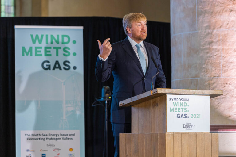 Het beeld toont een spreker in een donkerblauw pak met een lichtblauwe stropdas, die een presentatie geeft achter een houten spreekgestoelte. Op de achtergrond staat een banner met de tekst WIND. MEETS. GAS. en informatie over een symposium in 2021 georganiseerd door de New Energy Coalition. De setting lijkt formeel en professioneel, met thema's die waarschijnlijk gerelateerd zijn aan energie en duurzaamheid. Fotograaf: Jan Buwalda, Groningen.