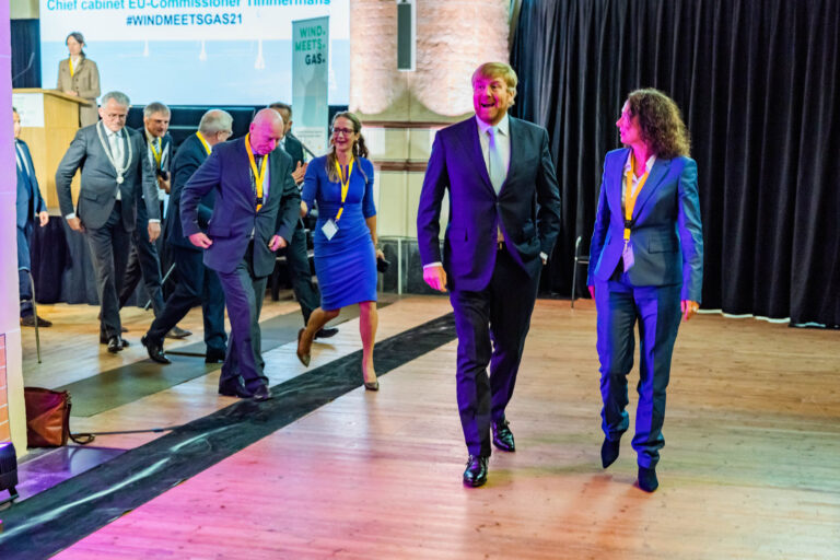 Dit beeld toont een groep mensen in formele kleding die over een houten vloer lopen tijdens een conferentie of evenement. Op de achtergrond is een podium zichtbaar met een sprekerspaneel en banners met teksten zoals #WINDMEETSGAS21. De omgeving lijkt professioneel en de personen dragen naamkaartjes om hun nek. De foto vangt een dynamische en vrolijke sfeer onder de deelnemers. Fotograaf: Jan Buwalda, Groningen.