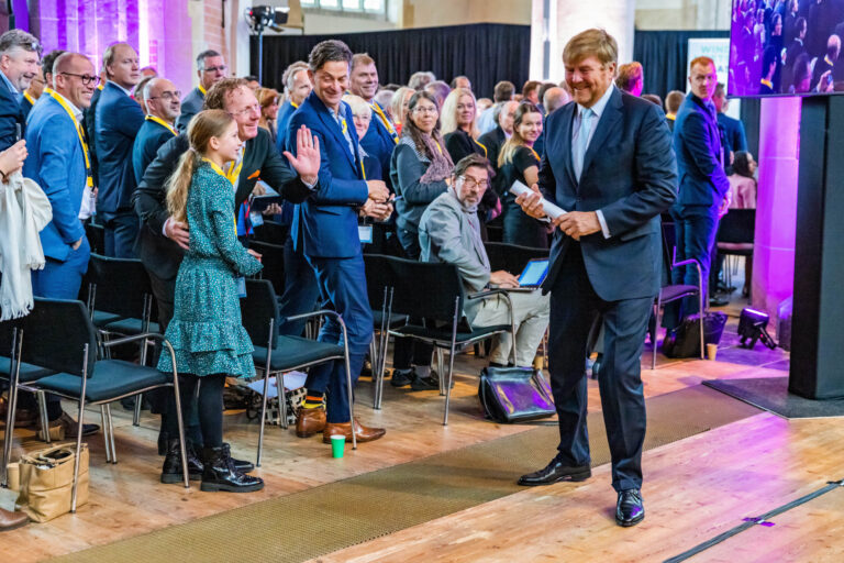 In deze kleurrijke foto genomen door Jan Buwalda, zien we een groep mensen in een zaal in Groningen. De sfeer is vrolijk en informeel. Een man in een donker pak, die vooraan staat, glimlacht terwijl hij met een papier in zijn hand staat. Mensen in het publiek lijken geïnteresseerd en sommigen lachen of zwaaien naar de man in het pak. De verlichting in de zaal voegt een warme, uitnodigende gloed toe aan de bijeenkomst.