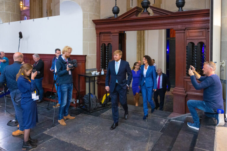 Dit is een foto genomen door Jan Buwalda in Groningen. Het beeld toont een formele gebeurtenis waarbij een aantal personen door een deuropening lopen, omringd door fotografen en cameramensen. De ruimte heeft een klassieke architectuur met stenen vloeren en houten elementen. De aanwezige mensen zijn netjes gekleed, passend bij de plechtigheid van de gelegenheid.