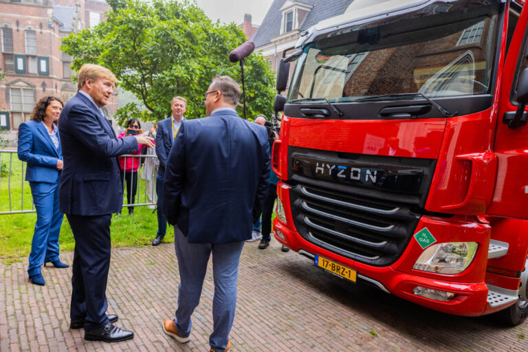 Een groep mensen is in gesprek voor een opvallende rode vrachtwagen met de naam HYZON op de grille. De foto is genomen in een buitenruimte met groene bomen en historische gebouwen op de achtergrond. De personen zijn netjes gekleed en lijken deel te nemen aan een officieel evenement. Fotograaf: Jan Buwalda, Groningen.