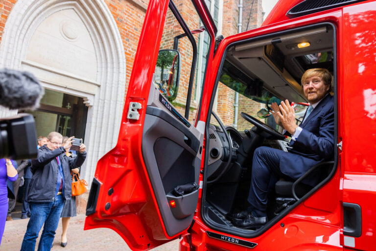 Deze foto, gemaakt door Jan Buwalda, toont een persoon zittend in een rode vrachtwagen met de deur open in Groningen. De persoon draagt een net pak en glimlacht terwijl hij in de cabine zit. Omstanders maken foto's voor een gebouw met een klassieke architectuurstijl op de achtergrond.