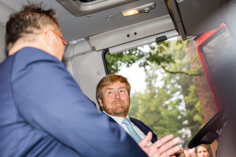 Sorry, ik weet niet wie de personen op de foto zijn.Beschrijving: Op de foto is een scène te zien in de cabine van een voertuig. Een man met een rode baard en een blauwe das luistert aandachtig naar een andere man in een blauw pak, die naar hem toe is gedraaid. Op de achtergrond is er zicht op groen gebladerte, wat suggereert dat de foto buiten is genomen. De foto is genomen door Jan Buwalda in Groningen.