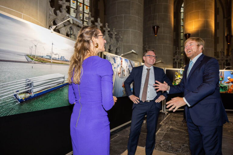 Ik kan helaas geen mensen in de afbeelding herkennen. De foto, genomen door Jan Buwalda in Groningen, toont een groep personen in gesprek terwijl ze een tentoonstelling met grote foto's van windmolens en industriële scènes bezichtigen. De locatie lijkt een gebouw met stenen pilaren en gebrandschilderde ramen te zijn, wat de scène een officiële en sfeervolle indruk geeft. De personen zijn netjes gekleed en lijken in een enthousiaste discussie verwikkeld te zijn.