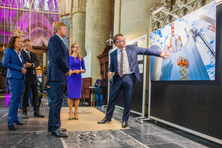 In de afbeelding zien we een groep mensen die aandachtig luisteren naar een spreker die wijst naar een groot foto paneel. Het paneel toont een industriële installatie met kranen en silo's tegen een helderblauwe lucht. De setting bevindt zich in een indrukwekkend gebouw met hoge gewelfde plafonds en kleurrijke glas-in-loodramen. De personen zijn gekleed in formele kleding, en de sfeer is professioneel en gefocust. Fotograaf: Jan Buwalda, Groningen.
