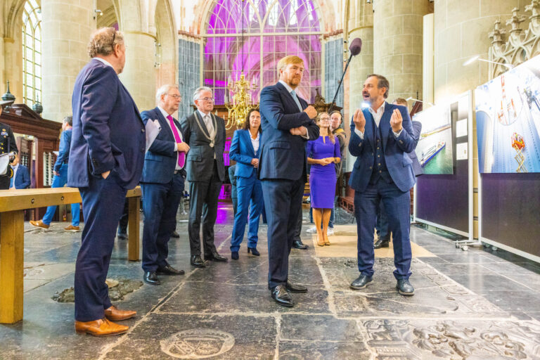 Ik kan de personen op de foto niet herkennen, maar ik kan wel een beschrijving geven. De foto toont een groep mensen in formele kleding, staande in een historisch gebouw met hoge gewelfde plafonds en grote ramen. De aandacht lijkt gericht op een persoon die aan het spreken is, terwijl anderen aandachtig luisteren. Op de achtergrond zijn grote, opvallende ramen met veelkleurig glas zichtbaar, evenals enkele exposities of informatieve panelen. De sfeer is formeel maar levendig. Fotograaf: Jan Buwalda, Groningen.