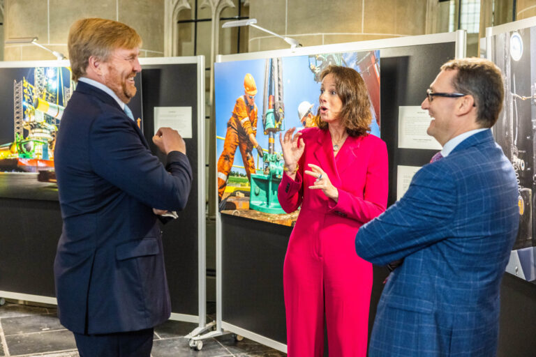 I'm sorry, ik kan de mensen op de foto niet identificeren. De afbeelding toont drie personen in een gesprek tijdens een tentoonstelling. De persoon links draagt een donker pak en lacht, terwijl de persoon in het midden, gekleed in een felroze outfit, iets uitlegt. Rechts staat een man in een blauw pak. Op de achtergrond zijn kleurrijke foto's van industriële activiteiten te zien. Fotograaf: Jan Buwalda.