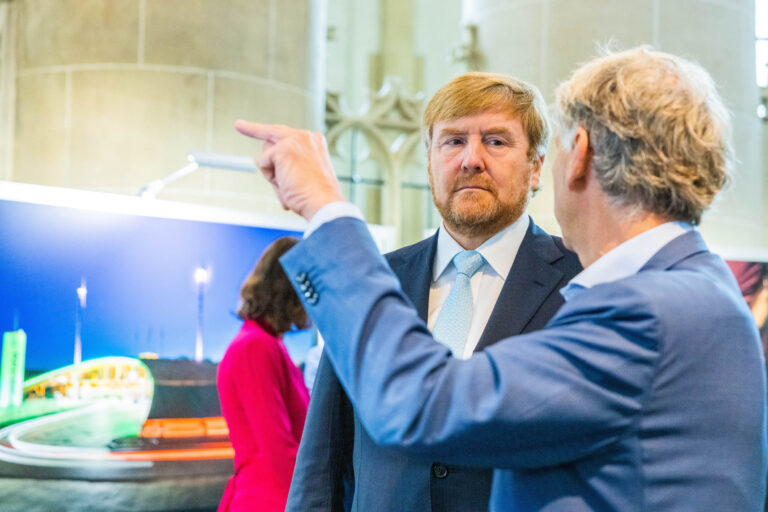 Het is een foto genomen door Jan Buwalda in Groningen. De foto toont een man die aandachtig luistert naar een andere man die naar iets lijkt te wijzen. De setting is waarschijnlijk binnen, met een kleurrijke afbeelding op de achtergrond en architectonische elementen die doen denken aan een tentoonstelling of presentatie.