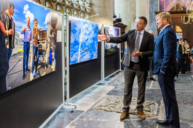 Je wordt getoond een expositieruimte in een imposant gebouw met hoge plafonds en kunstige pilaren. Twee mannen zijn in gesprek, waarbij één van de mannen wijst naar de tentoongestelde foto's. Deze foto's zijn kleurrijk en divers, en worden gepresenteerd op grote panelen. Op de achtergrond zijn meer mensen aanwezig, waarschijnlijk bezoekers van de expositie. Dit evenement lijkt plaats te vinden in een historisch gebouw, afgaand op de architectuur en de gedetailleerde versieringen.Fotograaf: Jan Buwalda, Groningen.