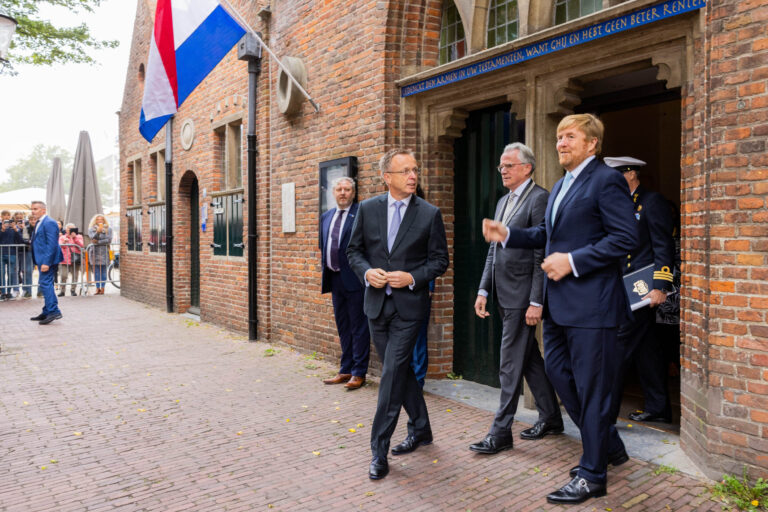 Het beeld toont een groep mannen in formele kleding die uit een historisch gebouw in Groningen komen. Boven de ingang van het gebouw is een opschrift zichtbaar. Aan de gevel hangt een Nederlandse vlag, en op de achtergrond staan mensen met camera's. De straat is bedekt met klinkers en de sfeer lijkt officieel en plechtig. Fotograaf: Jan Buwalda.