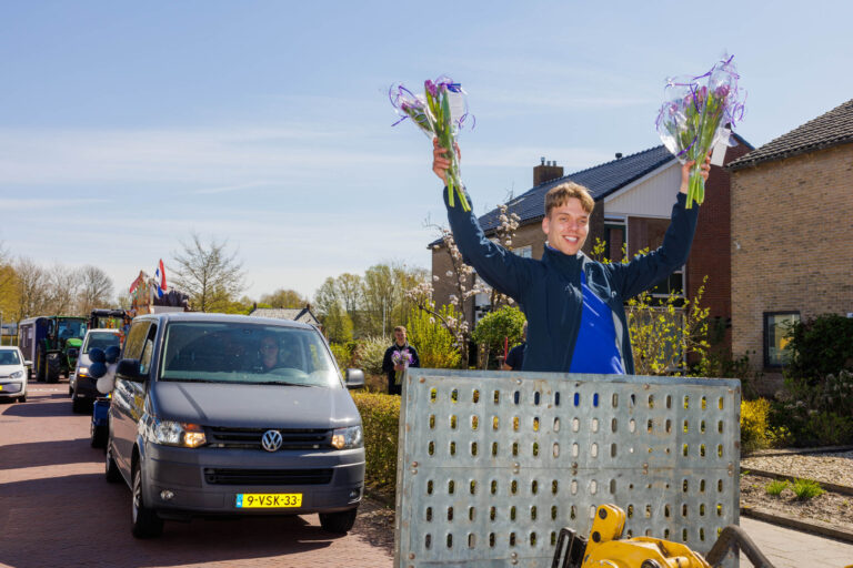 Een jonge man staat op een voertuig en houdt enthousiast twee boeketten met bloemen omhoog terwijl hij glimlacht. Op de achtergrond is een straat te zien met verschillende voertuigen, waaronder een Volkswagen busje en een tractor. Mensen langs de weg kijken toe, en er is een ontspannen en feestelijke sfeer. De zon schijnt helder op de scène, en er zijn enkele huizen en bomen op de achtergrond. Foto door Jan Buwalda, Groningen.