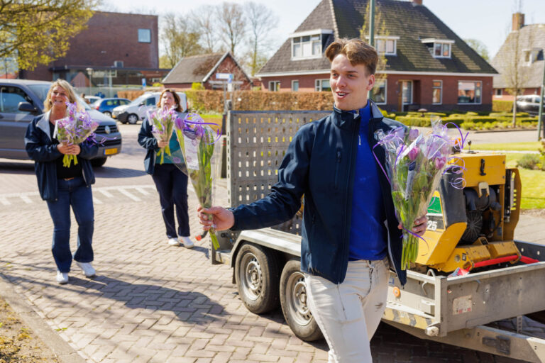 In deze foto, gemaakt door Jan Buwalda in Groningen, zien we drie mensen die op een zonnige dag bloemen uitdelen. Ze dragen blauwe jassen en lopen vrolijk over een straat met bakstenen. Op de achtergrond is een woonwijk met traditionele Nederlandse huizen zichtbaar. De bloemen zijn ingepakt in doorzichtige folie en met paarse linten versierd. Hun blije gezichten stralen een gevoel van gemeenschap en vreugde uit.