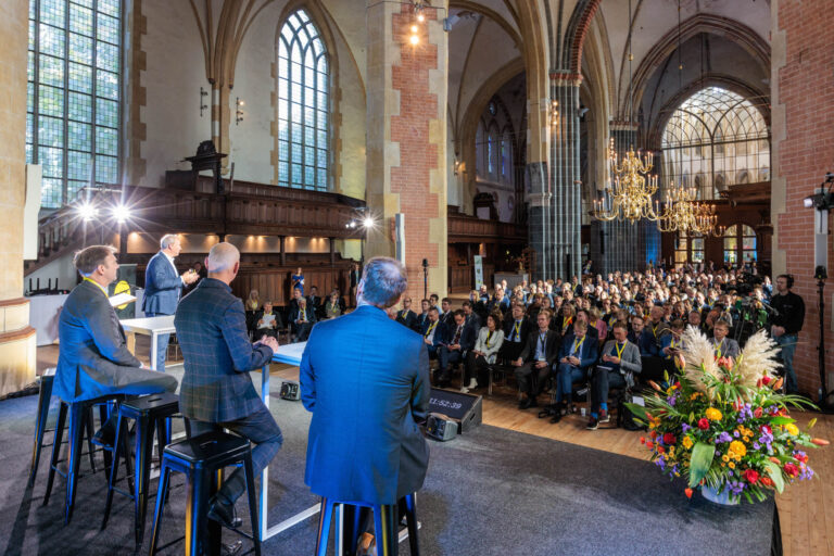 Dit beeld toont een levendig evenement binnen de muren van een indrukwekkende gotische kerk in Groningen. De ruimte is vol met aandachtige toehoorders die naar de sprekers op het podium luisteren. De hoge gewelven en grote glas-in-loodramen creëren een majestueuze achtergrond voor de bijeenkomst. Voor op de foto is een kleurrijk bloemstuk zichtbaar, dat bijdraagt aan de sfeer. De fotografie is verzorgd door Jan Buwalda.