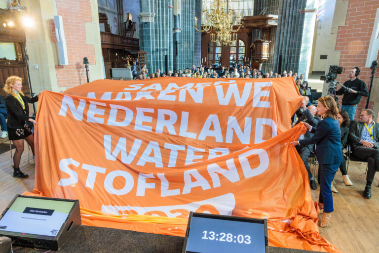 In een grote zaal, vermoedelijk een kerkgebouw, vindt een presentatie plaats waarbij een groot oranje doek met de tekst SAMEN MAKEN WE NEDERLAND WATERSTOFLAND wordt onthuld. Op de achtergrond zijn diverse toeschouwers te zien, terwijl het doek wordt vastgehouden door meerdere mensen. Er staat een monitor op de voorgrond die de naam Ulke Vermeulen toont en een tijdstip van 13:28:03. Op de achtergrond zijn de karakteristieke elementen van de kerk zichtbaar, inclusief hoge plafonds en grote ramen, wat een gevoel van grootsheid geeft aan het evenement. De foto is genomen door Jan Buwalda, in Groningen.