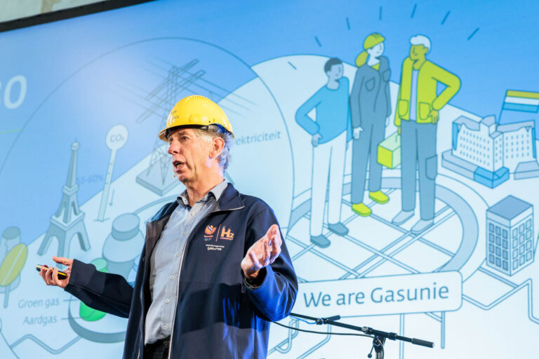 Sorry, ik kan niet vertellen wie er op de foto staat.In de afbeelding zie je een man met een veiligheidshelm die presenteert voor een groot scherm. Op het scherm is een illustratie te zien met verschillende elementen zoals gebouwen, mensen, en teksten zoals We are Gasunie. De man draagt een blauwe jas met het logo van WaterstofNet en Gasunie. De setting lijkt gericht op een presentatie of conferentie over energie of infrastructuur.Fotograaf: Jan Buwalda, Groningen.