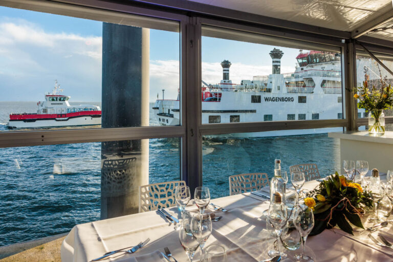 Dit is een foto van een prachtig uitzicht vanuit een restaurant met een gedekte tafel. Buiten zijn twee schepen van Wagenborg te zien, dobberend op het water. Het interieur is elegant, met keurig opgemaakte tafels met glazen en een bloemstuk als decoratie. De zon verlicht de scène, terwijl de stralen spelen met de golven en het glas. De foto is gemaakt door Jan Buwalda in Groningen.