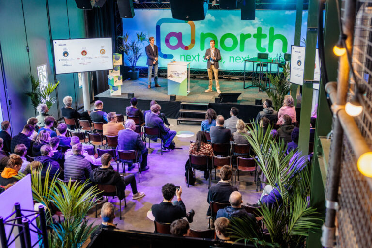 Dit beeld, vastgelegd door fotograaf Jan Buwalda in Groningen, toont een levendig zakelijk evenement waar een spreker op het podium een presentatie geeft aan een betrokken publiek. De zaal is modern ingericht met blauwe en groene lichteffecten, passend bij het thema van de bijeenkomst. Het aanwezige publiek zit aandachtig te luisteren, terwijl een groot scherm ter ondersteuning van de presentatie op de achtergrond zichtbaar is. Planten zijn strategisch geplaatst om de ruimte een frisse uitstraling te geven.