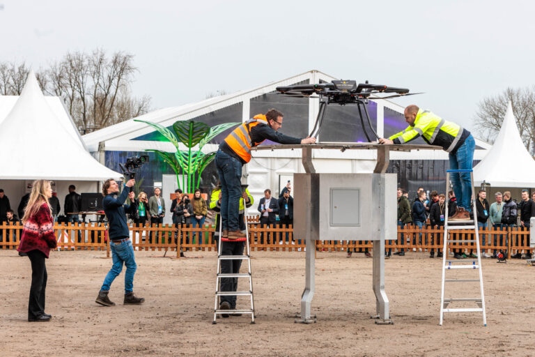 Op de foto, gemaakt door Jan Buwalda in Groningen, zien we een dynamisch tafereel op een buitenlocatie. Twee mannen in veiligheidskleding staan bovenop ladders en zijn bezig met het installeren of controleren van een grote drone die op een platform rust. In de achtergrond staan verschillende mensen te kijken, sommigen met camera's, wat erop lijkt te wijzen dat het om een evenement gaat. Er is een grote tent te zien en een houten hek die het gebied afschermt. De sfeer is bedrijvig en nieuwsgierigheid hangt in de lucht.