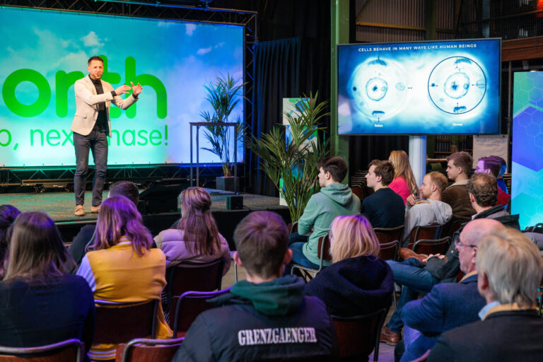 Een spreker staat op een podium voor een geïnteresseerd publiek tijdens een presentatie over cellen, met visuals op een groot scherm naast hem. De achtergrond is versierd met een groot paneel en enkele planten. De ruimte is modern en goed verlicht, met een sfeer van aandacht en interesse onder de aanwezigen.Fotograaf: Jan Buwalda, Groningen.