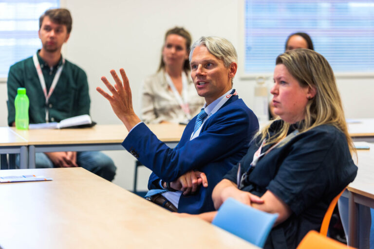 Een groep mensen zit aandachtig te luisteren in een klaslokaal. Een man in een blauw pak is aan het spreken en maakt een handgebaar terwijl anderen naar hem luisteren. De sfeer is die van een interactieve discussie of presentatie. De foto is helder en levendig, met een focus op de centrale figuren. Gemaakt door fotograaf Jan Buwalda in Groningen.