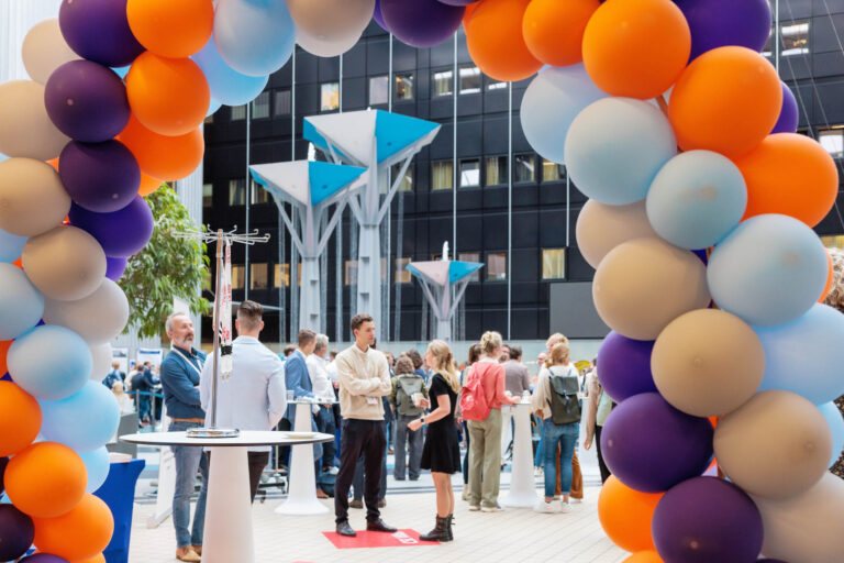 De foto, genomen door Jan Buwalda in Groningen, toont een levendige bijeenkomst in een modern gebouw. Op de voorgrond is een kleurrijke boog van ballonnen in tinten oranje, paars, en lichtblauw te zien. Onder deze boog zijn mensen gezellig in gesprek met elkaar. De achtergrond bevat architectonische elementen met strakke lijnen en symmetrische vormen, wat bijdraagt aan de moderne uitstraling van de omgeving.