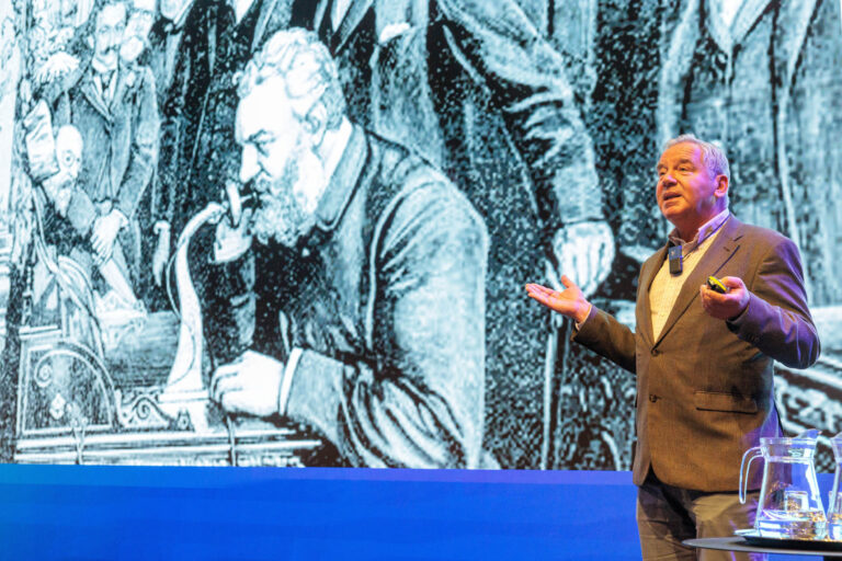 Dit beeld toont een man die een presentatie geeft op een podium. Op de achtergrond is een grote illustratie te zien van een historische scène met mensen die gebruikmaken van een ouderwetse telefoonachtige machine. De presentator draagt een pak en gebruikt een microfoon. Rechts op de voorgrond staat een tafel met een glazen karaf erop. De foto vangt een moment van interactie en uitleg, waarbij de spreker betrokken lijkt bij zijn verhaal. Fotograaf: Jan Buwalda, Groningen.