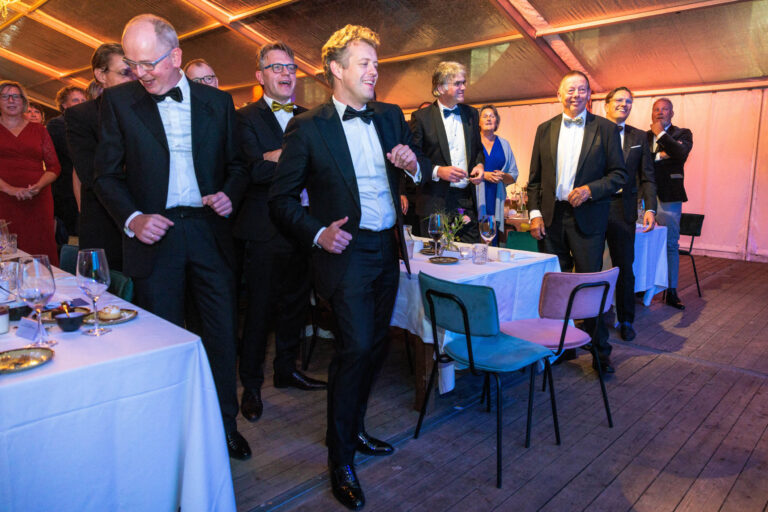 Een feestelijke bijeenkomst waarbij mensen in nette avondkleding staan en plezier hebben in een verlichte feesttent. De sfeer is informeel en vrolijk. Tafels zijn gedekt met glazen en borden, en de gasten lijken te genieten van het evenement. De foto is genomen door Jan Buwalda in Groningen.