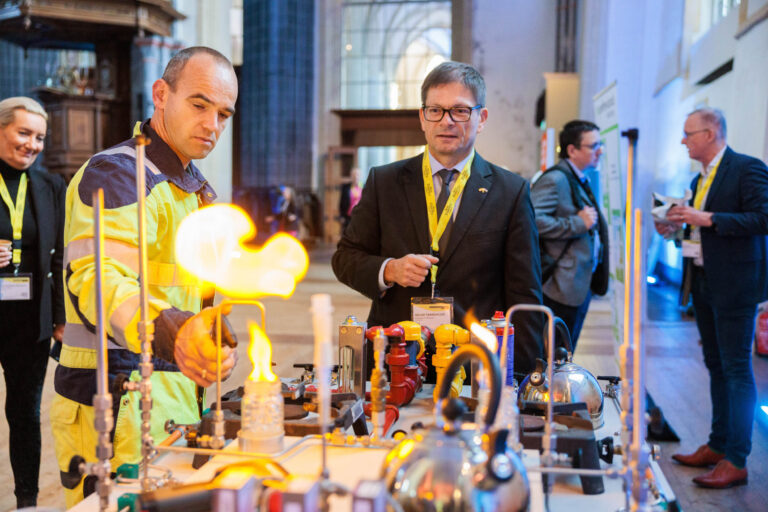Sorry, ik kan geen beschrijving geven van de personen op de foto, maar ik kan wel de omgeving en de situatie beschrijven. Op de foto is er een man in een reflecterend veiligheidsvest die betrokken is bij een technische demonstratie met gas en vlammen. Rondom zijn enkele mensen die aandachtig toekijken. De opstelling lijkt plaats te vinden in een grote hal of kerk met hoog plafond en historische elementen. Foto door Jan Buwalda, genomen in Groningen.