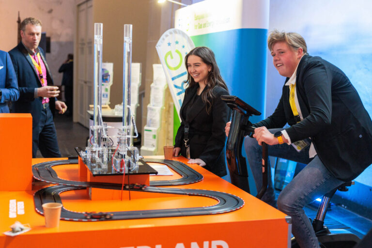 Een groep mensen is bezig met een interactieve opstelling waarbij een persoon op een hometrainer zit en een modelracebaan lijkt te bedienen. Op de achtergrond zijn promotiematerialen te zien. De sfeer is levendig en plezierig. Foto door Jan Buwalda, genomen in Groningen.