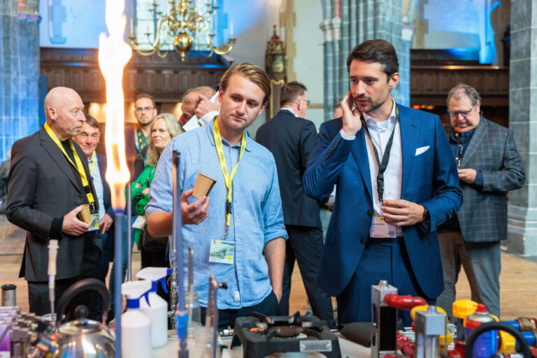 Ik kan de personen op de foto niet herkennen, maar ik zal een beschrijving geven:Op de foto is een groep mensen te zien die deelnemen aan een evenement binnen een historische zaal. De ruimte heeft hoge stenen zuilen en een grote kroonluchter aan het plafond. Voorop in het beeld staan twee mannen die lijken te discussiëren over een technische opstelling met diverse wetenschappelijke instrumenten en een vlam. Andere mensen staan op de achtergrond te praten en te observeren. Iedereen draagt gele naambadges. De sfeer lijkt professioneel en geïnteresseerd. Fotografie door Jan Buwalda in Groningen.