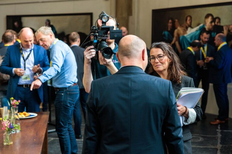 Deze foto toont een dynamische scène op een professionele bijeenkomst in Groningen. Mensen zijn bezig met gesprekken en interacties. Een man met een camera legt momenten vast, terwijl anderen schijnbaar in discussie zijn of netwerken. In de achtergrond zijn er meer mensen in gesprek, en er hangt kunst aan de muur. Het geheel straalt een sfeer van professionele samenwerking en uitwisseling van ideeën uit. Fotograaf: Jan Buwalda.