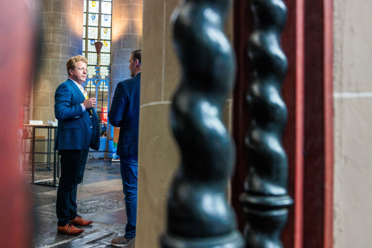 De foto toont twee mannen in gesprek in een ruimte met hoge stenen pilaren en glas-in-lood ramen, wat lijkt op een kerk of monumentaal gebouw. Ze dragen nette pakken en staan tegenover elkaar. De scène is vastgelegd vanuit een hoek waarin een houten en donker metalen element op de voorgrond zichtbaar is. De sfeer is formeel en intiem tegelijk. Fotograaf: Jan Buwalda, Groningen.