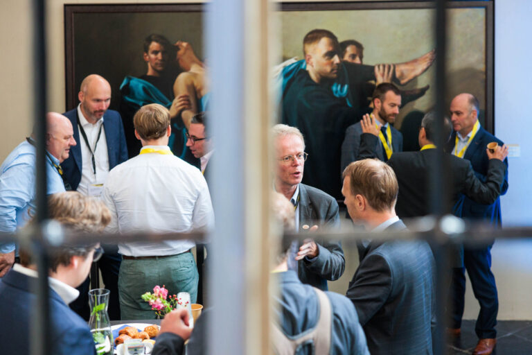 Het beeld toont een bijeenkomst waarbij mensen in nette kleding met elkaar in gesprek zijn. Op de achtergrond hangt een groot schilderij met een klassiek thema. De setting oogt professioneel, met deelnemers die badges dragen, mogelijk voor een conferentie of netwerkbijeenkomst. Er is een tafel met hapjes en drankjes te zien, wat bijdraagt aan de ontspannen sfeer. Deze foto is genomen door Jan Buwalda in Groningen.