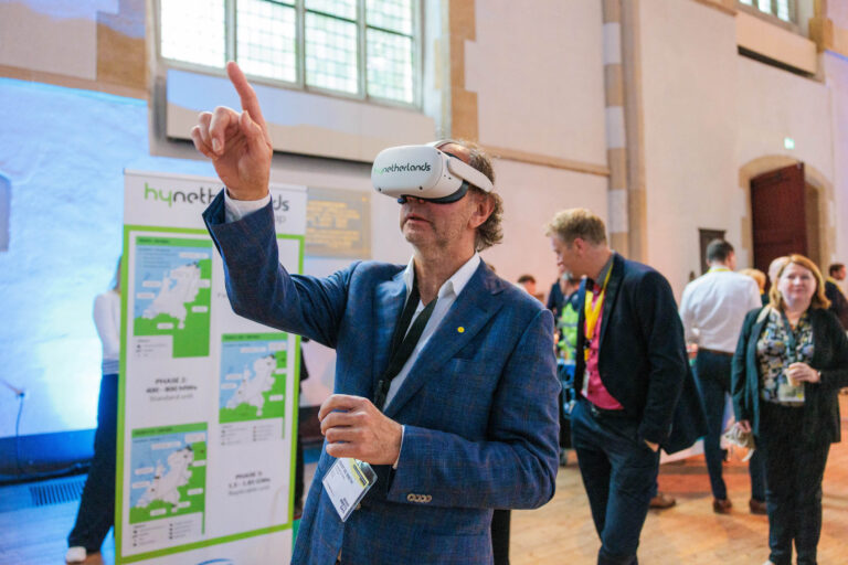 Een man in een blauw pak staat in een grote zaal en verwendet een virtual reality-bril. Hij wijst met zijn vinger naar iets in de virtuele omgeving. Op de achtergrond zijn meerdere mensen te zien die zich in de ruimte bewegen. Er is een banner met informatie over hynetherlands zichtbaar. De ruimte heeft grote ramen en een houten vloer. Foto door Jan Buwalda in Groningen.